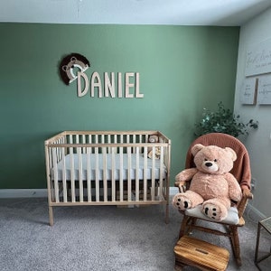 May include: A nursery with a wooden crib, a rocking chair with a teddy bear, and a wooden name sign that says "DANIEL" on a sage green wall.