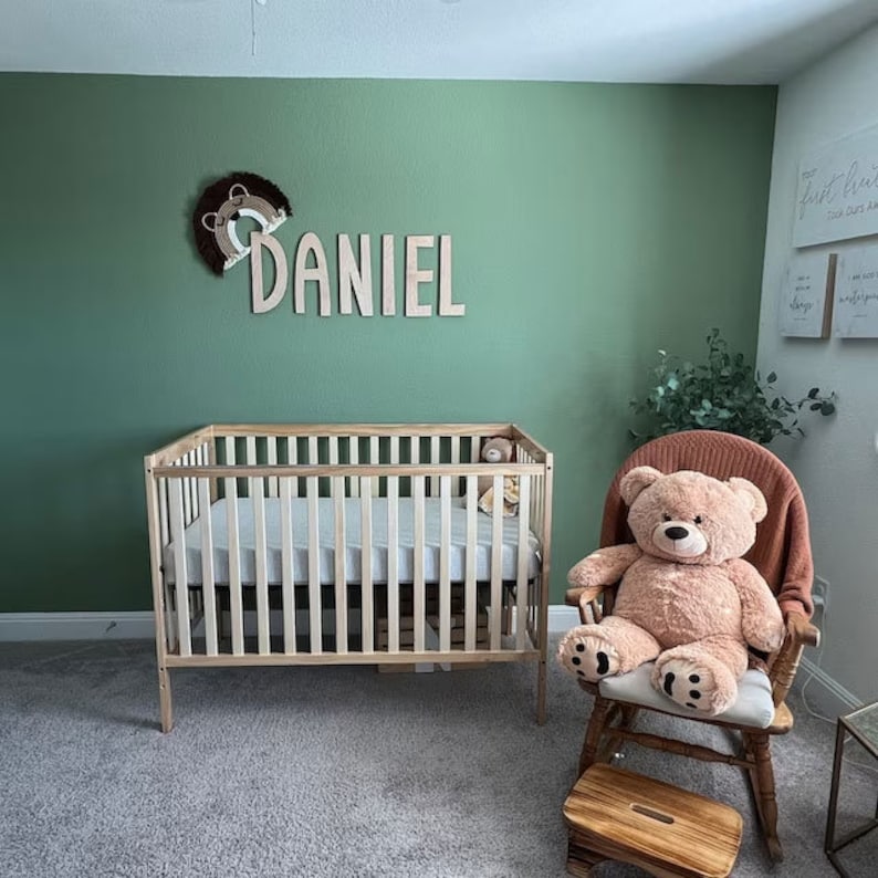 May include: A nursery with a wooden crib, a rocking chair with a teddy bear, and a wooden name sign that says "DANIEL" on a sage green wall.