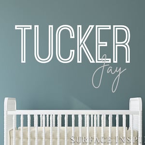 May include: White crib with a white wall behind it. The wall has a white vinyl decal with the name "TUCKER Jay" in a modern font.