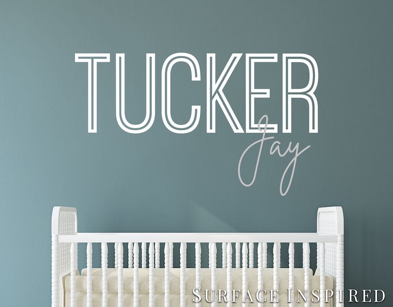 May include: White crib with a white wall behind it. The wall has a white vinyl decal with the name "TUCKER Jay" in a modern font.