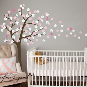 May include: A white crib with a pink and white patterned mattress and a brown tree with pink flowers and pink butterflies on a gray wall. The tree is a wall decal.