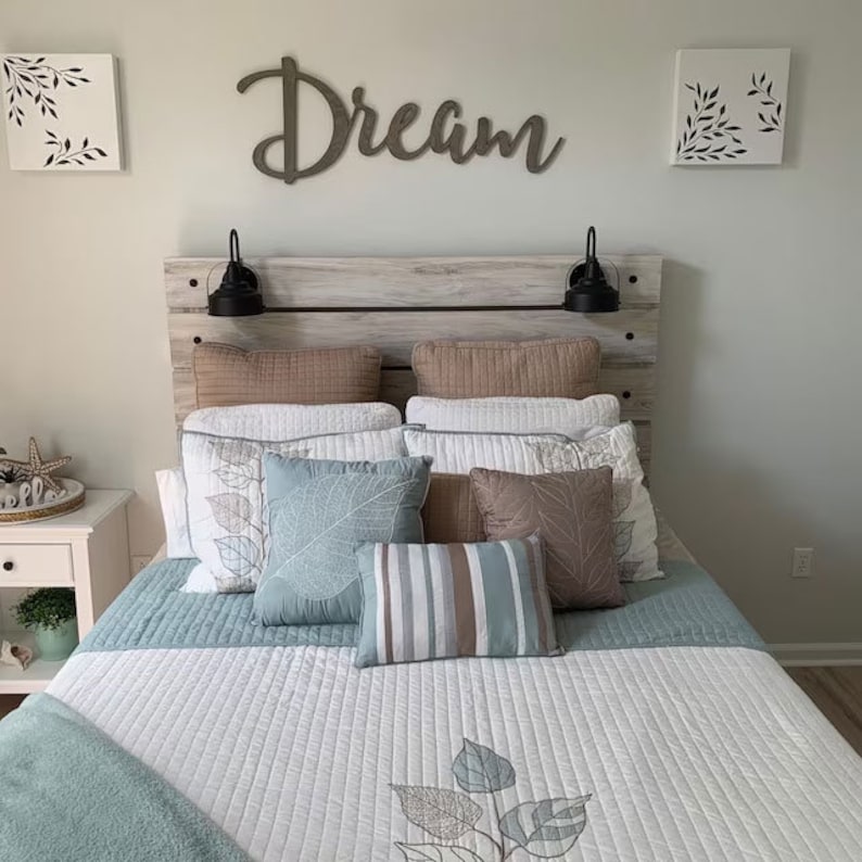 May include: A white bed with a wooden headboard and a blue and white quilt. The headboard has a sign that says "Dream" above it. There are several pillows on the bed, including a blue pillow with a leaf pattern, a striped pillow, and a brown pillow with a leaf pattern.