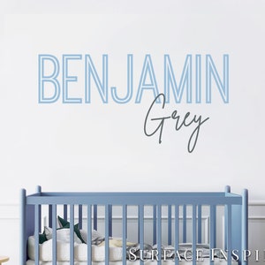 May include: A light blue crib with a white mattress and a blue and white patterned blanket. The wall behind the crib has a blue and white name decal that reads "BENJAMIN Grey".