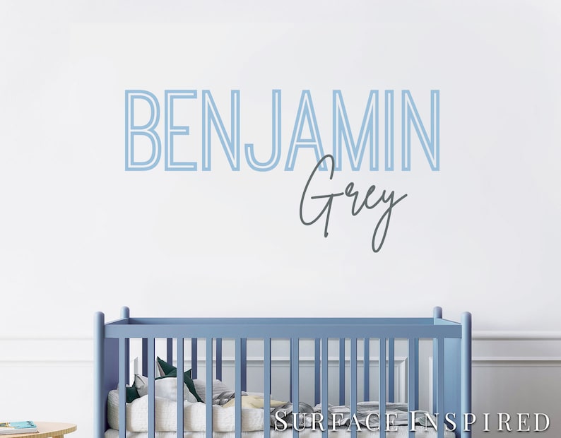 May include: A light blue crib with a white mattress and a blue and white patterned blanket. The wall behind the crib has a blue and white name decal that reads "BENJAMIN Grey".