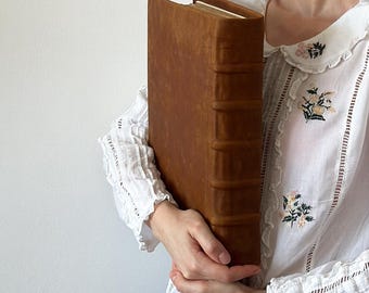 May include: A large, brown leather-bound book with decorative stitching and raised edges. The book is being held by a person.