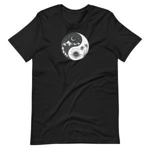 Puede incluir: Una camiseta negra de manga corta con un gráfico circular en el pecho. El diseño es un símbolo de yin-yang dividido en dos mitades: un lado representa una escena nocturna con montañas, árboles y una luna creciente, mientras el otro muestra una playa.