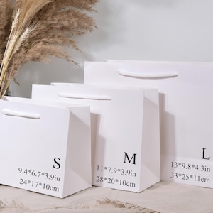 May include: Three white paper gift bags of varying sizes, labeled S, M, and L. Each bag has a white handle and printed dimensions in inches and centimeters. The bags are arranged in a row, with dried plants in the background.