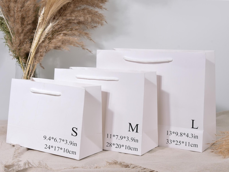 May include: Three white paper gift bags of varying sizes, labeled S, M, and L. Each bag has a white handle and printed dimensions in inches and centimeters. The bags are arranged in a row, with dried plants in the background.