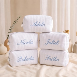 May include: Five white rectangular cosmetic bags with a waffle-textured fabric. Each bag features a silver zip with a round pull and is embroidered with a unique name in light blue script: Adele, Nicola, Juliet, Roberta, and Faithe.