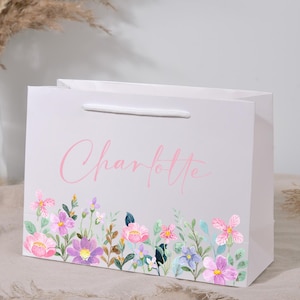 May include: A white gift bag with a floral design at the bottom and the name "Charlotte" in pink script. The floral design features pink, purple, and blue flowers with green leaves. The bag has a white handle.