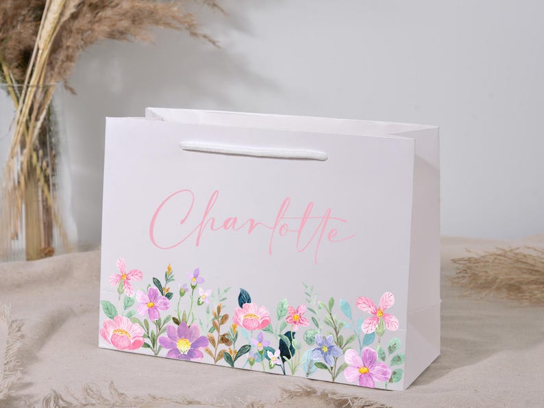 May include: A white gift bag with a floral design at the bottom and the name "Charlotte" in pink script. The floral design features pink, purple, and blue flowers with green leaves. The bag has a white handle.