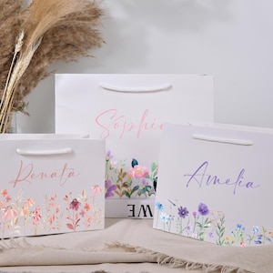 May include: Three white gift bags with floral designs and names in cursive: Renata, Sophie, and Amelia. Each bag has a different floral border and a matching name in a different color.