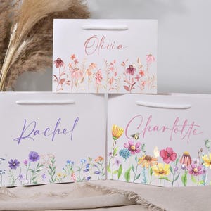 May include: Three white gift bags with floral designs and names: "Olivia" in a light brown script, "Rachel" in purple, and "Charlotte" in pink. Each bag features a different watercolor floral border with various flowers and greenery.