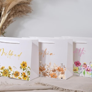 May include: White gift bags with floral designs and names in gold and pink script. The bags feature sunflowers, wildflowers, and other botanical illustrations. Each bag has a white handle.