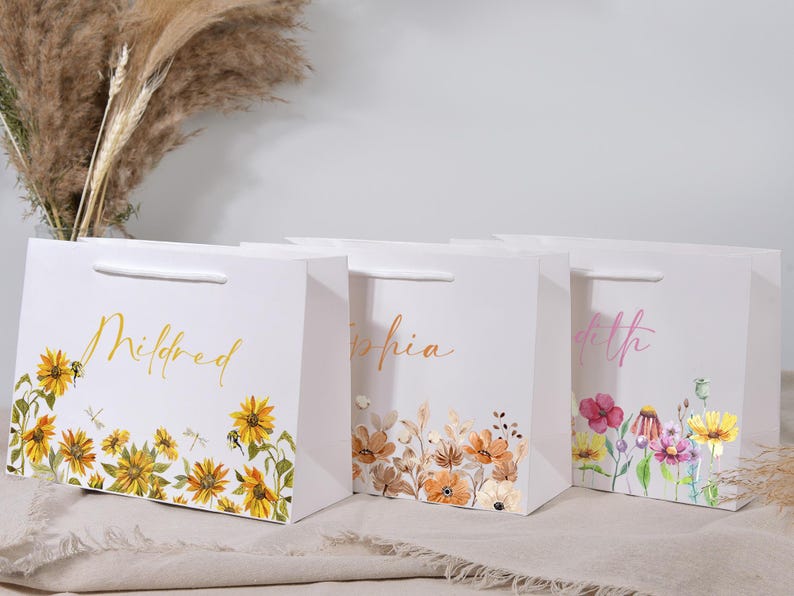 May include: White gift bags with floral designs and names in gold and pink script. The bags feature sunflowers, wildflowers, and other botanical illustrations. Each bag has a white handle.
