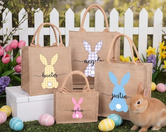 May include: A collection of burlap tote bags with bunny designs in yellow, purple, pink, and blue. Each bag features a different name: Hunter, Angela, Sophia, and Justin. The bags have tan handles and are set on a bed of green grass with Easter eggs and a rabbit.
