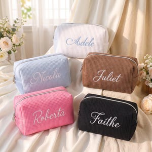 May include: Five rectangular waffle-textured cosmetic bags with silver zippers. The bags are white with blue embroidery reading Adele, light blue with white embroidery reading Nicola, brown with white embroidery reading Juliet, pink with white embroidery reading Roberta, and black with white embroidery reading Faithe.