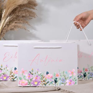 May include: Three white gift bags with floral watercolor designs and names in pink script. The bags have a handle and a colorful floral border with pink, purple, and blue flowers. One bag is being held by a hand.