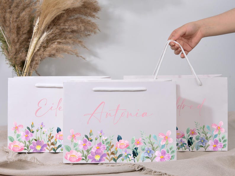 May include: Three white gift bags with floral watercolor designs and names in pink script. The bags have a handle and a colorful floral border with pink, purple, and blue flowers. One bag is being held by a hand.