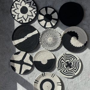 May include: Nine circular woven wall baskets in black and white geometric patterns. The designs include stripes, spirals, and radial motifs. The baskets are displayed on a grey surface. The word "SUZZI" is printed at the bottom of the image.