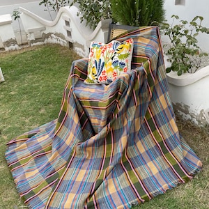 Könnte beinhalten: Eine karierte Decke in Gold-, Blau-, Grün- und Rosatönen ist über einen Stuhl drapiert. Ein dekoratives Kissen mit floralem Design liegt auf dem Stuhl. Die Decke hat einen Fransenrand und liegt auf einer Rasenfläche.