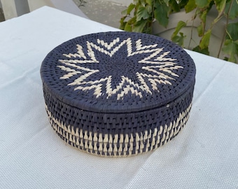 May include: A round, woven basket with a lid. The basket is navy blue with a starburst design on the lid in natural beige. The sides of the basket are also navy blue and beige. The basket is sitting on a white surface.