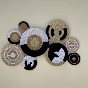 May include: A collection of woven wall decor in various sizes and patterns. The circular pieces feature combinations of black, white, and tan rope, creating a modern, geometric design. The arrangement is displayed against a neutral wall.
