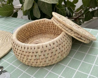 May include: A round, woven basket with a matching lid, crafted from light-colored natural fibers. The basket is open, revealing its interior, and sits on a green and white checkered tablecloth. A woven coaster is visible to the left.