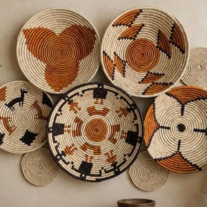 May include: A collection of seven round woven wall baskets in varying sizes. The baskets feature coiled designs in cream, burnt orange, and black, with patterns including geometric shapes, stylized animal figures, and human-like silhouettes.