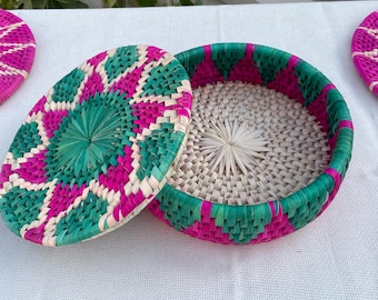 May include: A woven basket with a lid, featuring a vibrant design in green, pink, and white. The lid is oval-shaped, and the basket's interior is visible. The basket is likely handmade and suitable for storage or decoration.