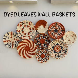 Puede incluir: Una colección de nueve cestas de pared tejidas en varios tamaños y patrones. Las cestas presentan fibras teñidas naturales, rojas y negras, creando diseños geométricos y en espiral. El texto "DYED LEAVES WALL BASKETS" se muestra arriba.
