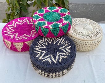 May include: Four round woven storage baskets with lids. The baskets are in shades of pink, green, blue, and natural beige. Each lid features a starburst or geometric design. The baskets are made of natural materials.