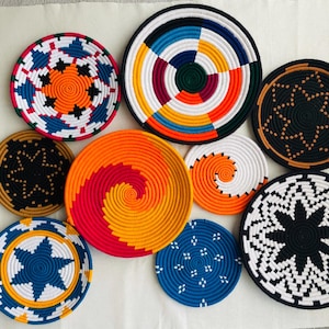 May include: A set of nine colorful woven wall decor plates. The plates are round and feature various geometric patterns in shades of blue, red, orange, yellow, black, and white.