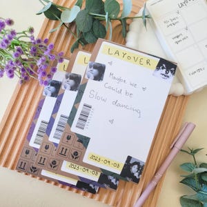 Puede incluir: Una pila de blocs de notas blancos y marrones con la palabra "LAYOVER" y el texto "Maybe we Could be Slow dancing". Cada bloc tiene una foto y un código de barras. Un bolígrafo rosa y flores moradas también están en la imagen.