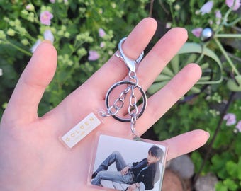 May include: A clear acrylic keychain with a silver metal clasp and chain. The keychain features a photograph of a person in a black jacket and jeans. A small rectangular tag with the word "GOLDEN" is also attached.