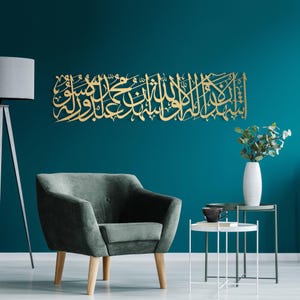 Könnte beinhalten: Ein goldfarbenes, verziertes arabisches Kalligraphie-Wandkunstwerk ist an einer türkisblauen Wand angebracht. Das Kunstwerk ist rechteckig und zeigt eine fließende Schrift. Ein grauer Sessel, Beistelltische und eine weiße Vase mit Grünzeug befinden sich im Vordergrund.