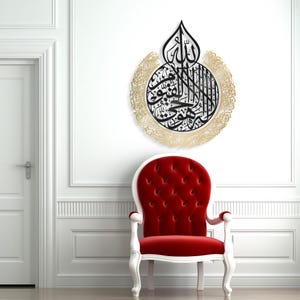 May include: A black and gold metal wall art featuring Islamic calligraphy in a circular design. The calligraphy is surrounded by a decorative border. The art is mounted on a white wall.