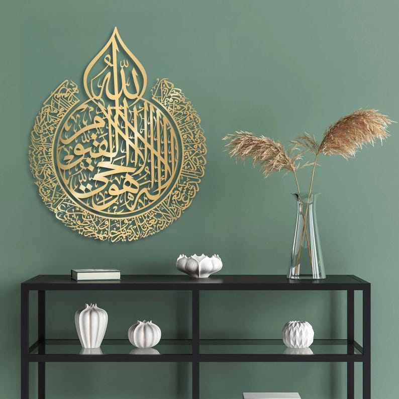 May include: Gold metal wall art featuring intricate Arabic calligraphy in a circular design. The art is displayed on a green wall above a black shelf with decorative white ceramic vases.