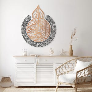 May include: A decorative wall art piece featuring intricate Arabic calligraphy in black and copper tones. The calligraphy is arranged in a circular pattern with a central design resembling a teardrop.