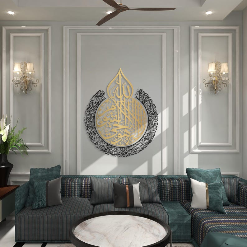 May include: A gold and black metal wall art piece with Arabic calligraphy in a circular design. The art is hanging on a white wall in a living room setting with a sofa and coffee table.