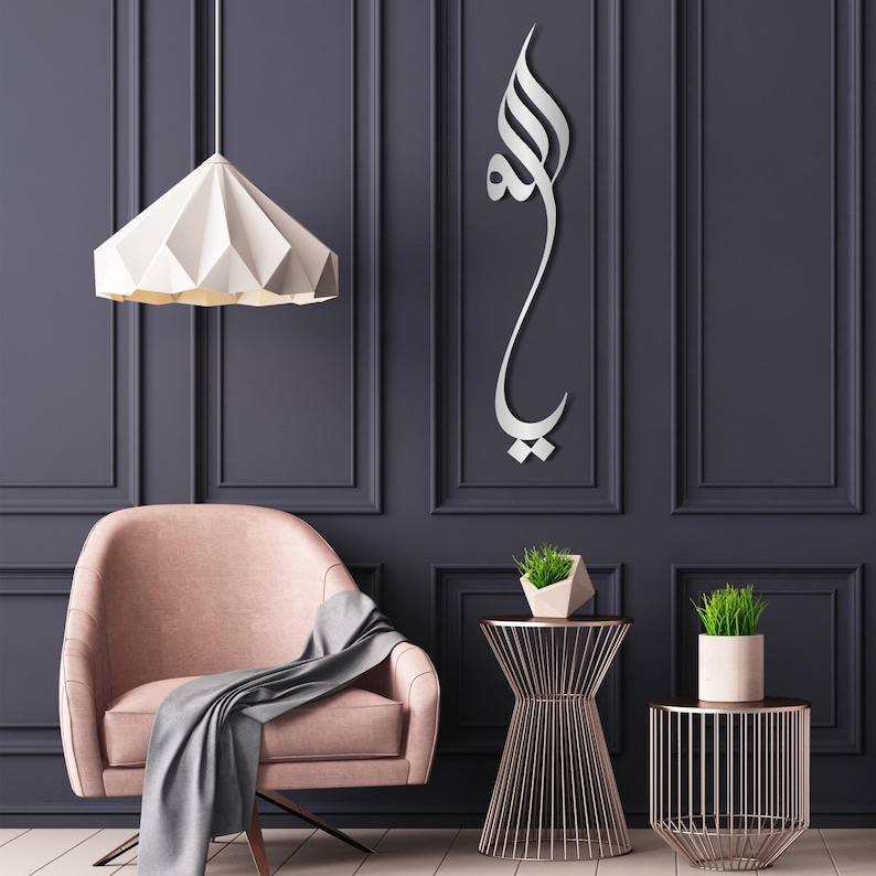 May include: A vertical silver-colored wall art piece featuring Arabic calligraphy script. The metallic design is mounted on a dark paneled wall, displaying a flowing, elongated aesthetic with distinct characters that form a singular, cohesive vertical composition.