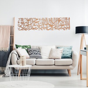 May include: A large, ornate, copper-colored metal wall art piece featuring Arabic calligraphy. The artwork is mounted on a white wall above a beige sofa with decorative pillows. The room is decorated in a modern style.