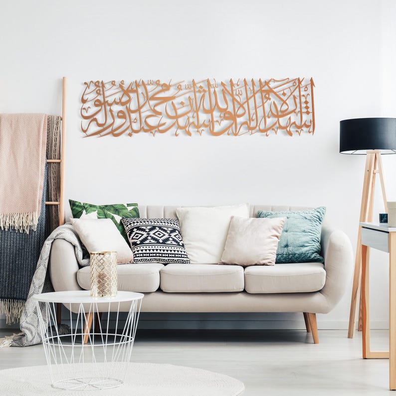 May include: A large, ornate, copper-colored metal wall art piece featuring Arabic calligraphy. The artwork is mounted on a white wall above a beige sofa with decorative pillows. The room is decorated in a modern style.