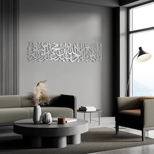 May include: A large, white, three-dimensional Arabic calligraphy art piece hangs on a gray wall. The artwork is in a rectangular shape and features intricate script. The room has modern furniture, including a gray sofa, a round coffee table, and a black floor lamp.