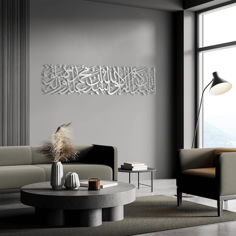 May include: A large, white, three-dimensional Arabic calligraphy art piece hangs on a gray wall. The artwork is in a rectangular shape and features intricate script. The room has modern furniture, including a gray sofa, a round coffee table, and a black floor lamp.