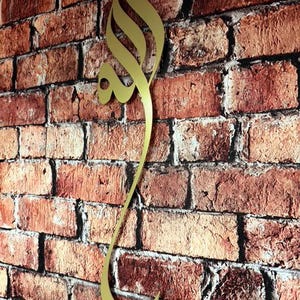 May include: A vertical gold-colored metal wall art piece featuring elegant Arabic calligraphy. The design is elongated and fluid, showcasing intricate curves and sharp points that form a continuous script. The metallic surface has a smooth finish.