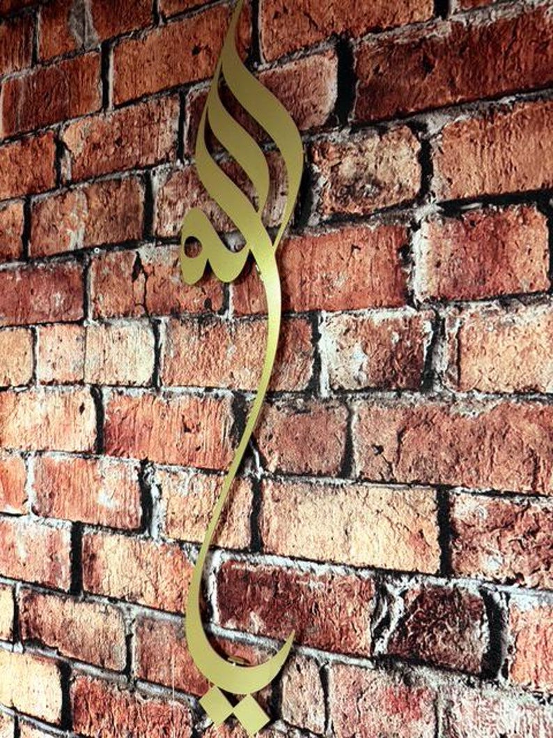 May include: A vertical gold-colored metal wall art piece featuring elegant Arabic calligraphy. The design is elongated and fluid, showcasing intricate curves and sharp points that form a continuous script. The metallic surface has a smooth finish.