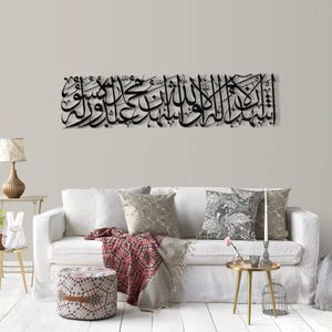 May include: Black metal wall art featuring Arabic calligraphy, mounted above a white sofa. The artwork is a long, horizontal piece with intricate script. The room includes a white sofa, patterned pillows, and a small side table with a lamp.