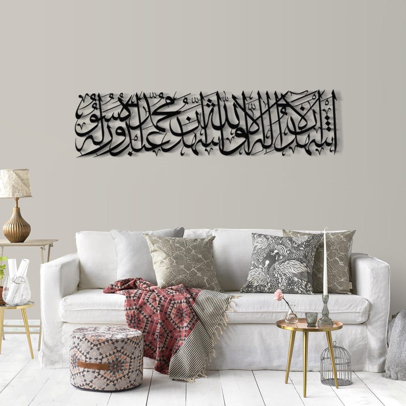 May include: Black metal wall art featuring Arabic calligraphy, mounted above a white sofa. The artwork is a long, horizontal piece with intricate script. The room includes a white sofa, patterned pillows, and a small side table with a lamp.