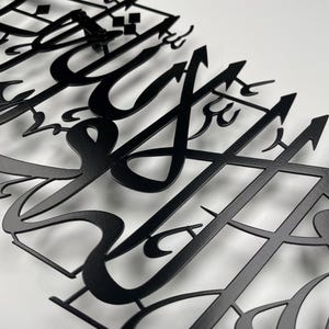 May include: Close-up of a black metal wall art piece featuring intricate Arabic calligraphy. The artwork is cut from metal and mounted on a white surface, showcasing the detailed design and elegant script. The piece is a decorative item.